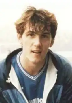 A brown-haired man in a blue football shirt with a blue tracksuit top over it, smiling slightly