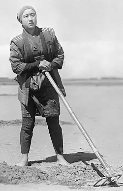 Black and white photograph of Akiko Kazami barefoot with a hoe and standing next to soil that has been dug up