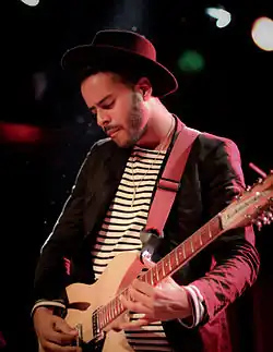 George William Lewis Jr., aka Twin Shadow, wearing a horizontal striped shirt under a dark blazer and a fedora hat, playing electric guitar onstage