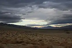 A landscape shot of a long, dry valley. The sky is partially clouded over but blue sky breaks through in patches. It is a showcase of Nevada's natural beauty.