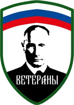 The Logo and patch of the Redut formation "Veterans", with the Russian tricolour at the top horizontally, the edges being green and the below part with white background and a black portrait of Putin in the middle, with the text "Veterany" or "Veterans" written below him.