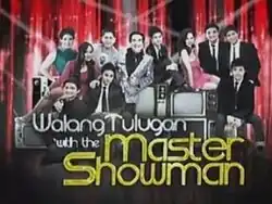 An image of the cast of Walang Tulugan with the Master Showman, with televisions surrounding them, and a red curtain on their back. The show title is displayed on the lower part of the image.