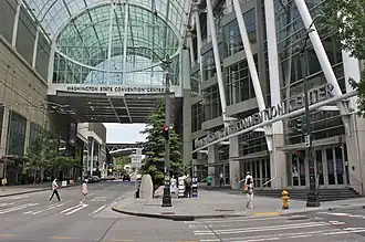 A street under a tall, arched glass bridge, as seen from a sidewalk