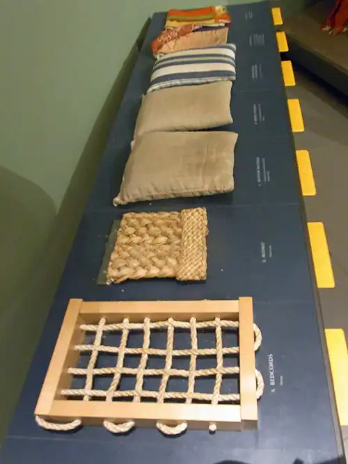 Touchable museum samples illustrating a 1590s bed: the bedcords, plaited-rush[6] bedmat, a flockbed and a featherbed in dun ticking, a downbed in striped ticking, and the bedlinen[5]