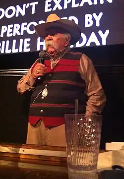An elderly white male with a long white mustache. He is wearing a cowboy hat and striped waistcoat while holding a microphone. He is standing in front of a screen.