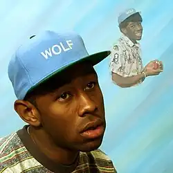 A close-up portrait of Tyler wearing a light blue baseball cap with "WOLF" embroidered in white letters, with a vacant and unfocused expression, against a light blue background consisting of blue, white and green blurred stripes. In the upper right corner, there is a smaller, faded image of Tyler cut off at the waist, cupping an inhaler in his hands, wearing a patterned shirt and the same blue cap, with "GOLF" embroidered on it.