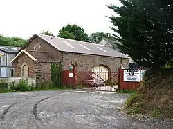 A lane, cut into the ground on the right, leads to a red fence and large single-storey stone building