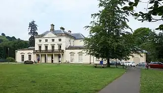 The front of Plas Machynlleth, a three-storey Georgian manor house with a cream-coloured exterior. A portico spanning the width of the main house is held up by five pillars, and there are single-storey wings on both sides of the building.