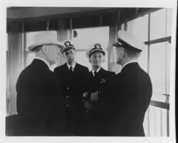 Black and white photograph of two men in naval uniforms facing the camera talking to two men, one in naval uniform and one wearing a suit and hat, both standing with their backs to the camera